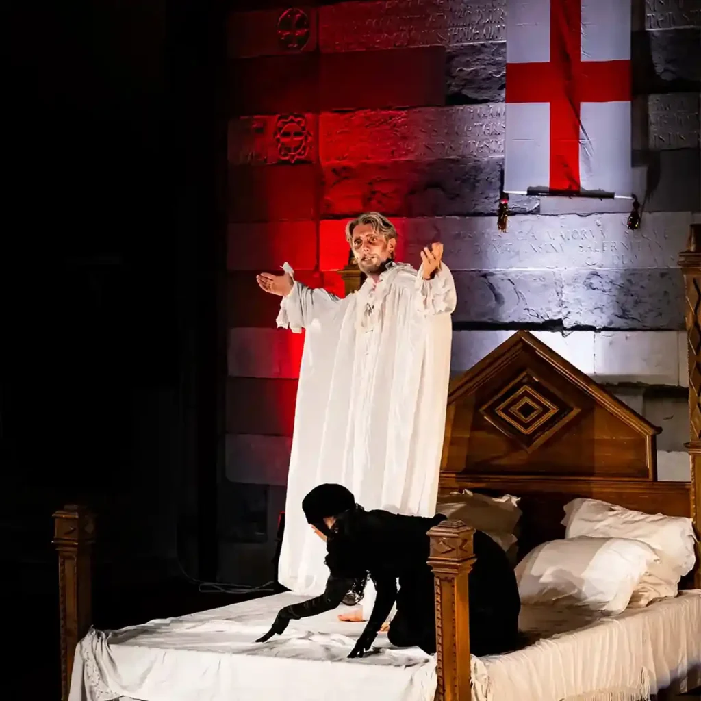 Attori in scena durante uno spettacolo teatrale con luci rosse e bandiera di San Giorgio sullo sfondo