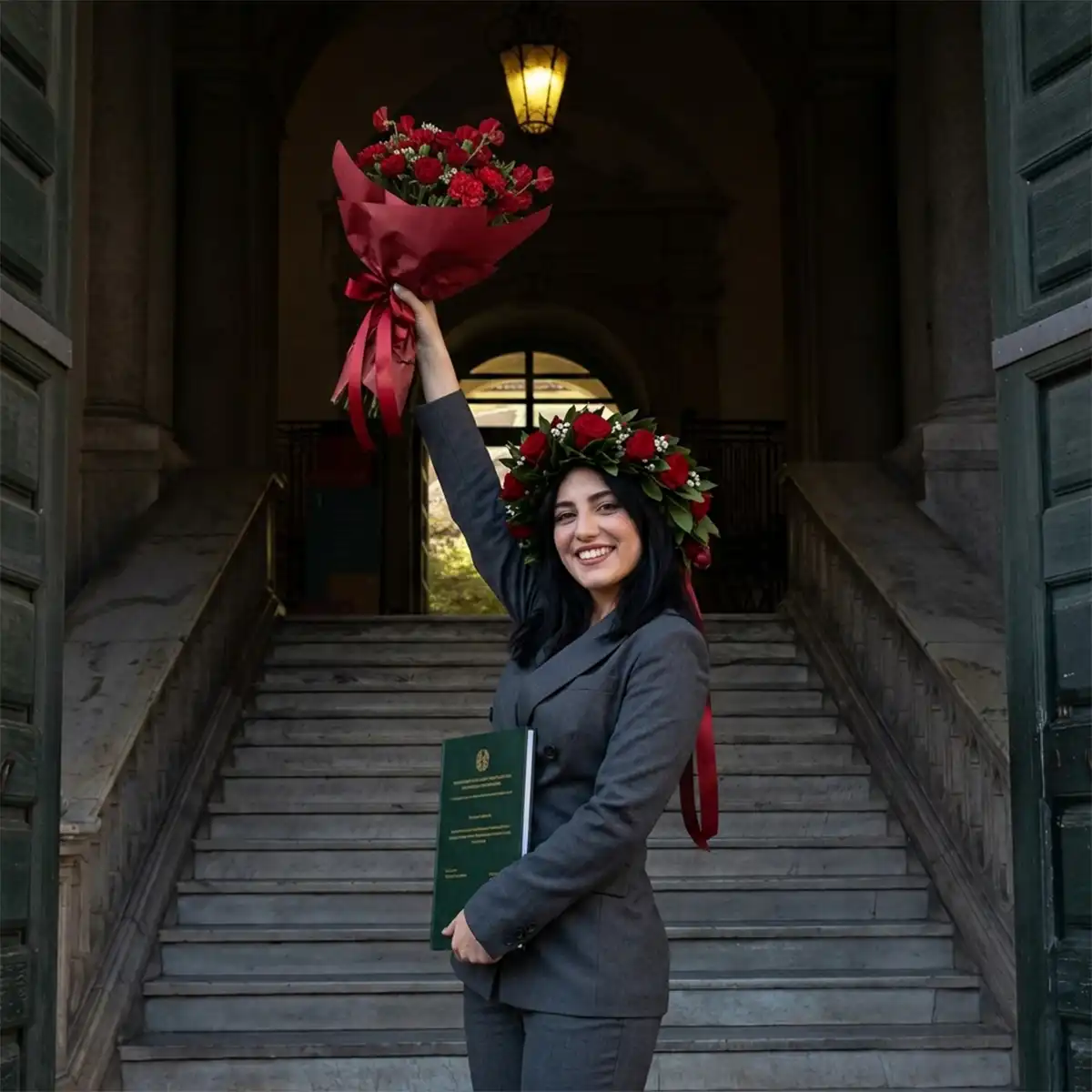Neolaureata con corona d'alloro alza un bouquet di rose rosse sulla scalinata dell'università