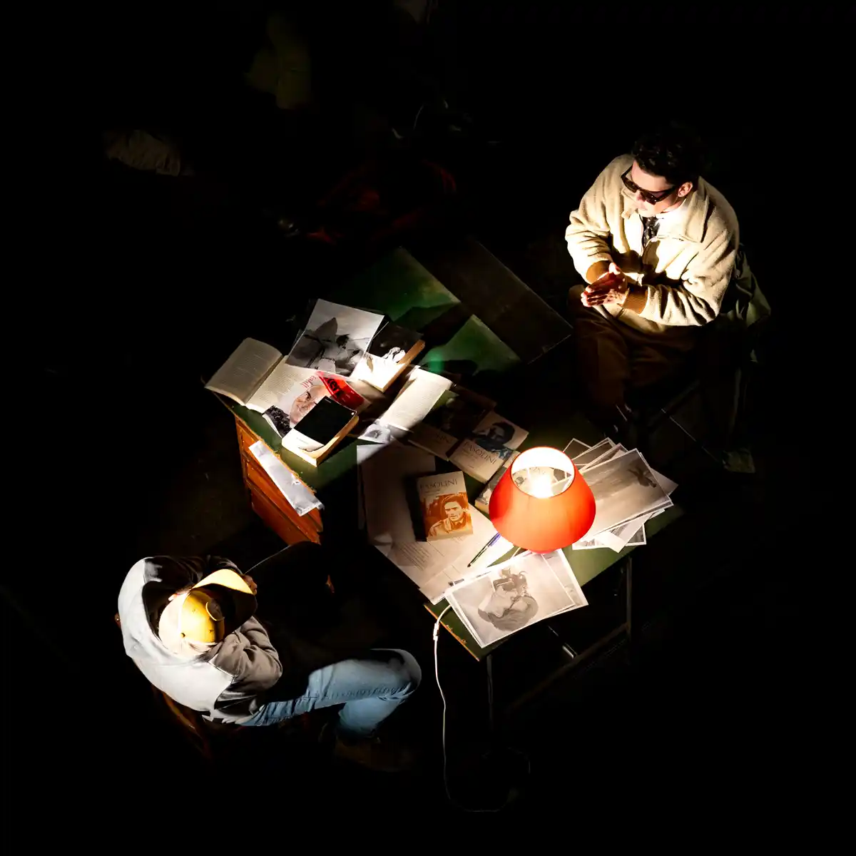 Due attori attorno a un tavolo con libri e fotografie illuminato da una lampada rossa, scena teatrale ripresa dall'alto