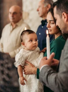 Bambino in abito bianco in braccio ai genitori accanto alla candela accesa durante il battesimo a Portofino